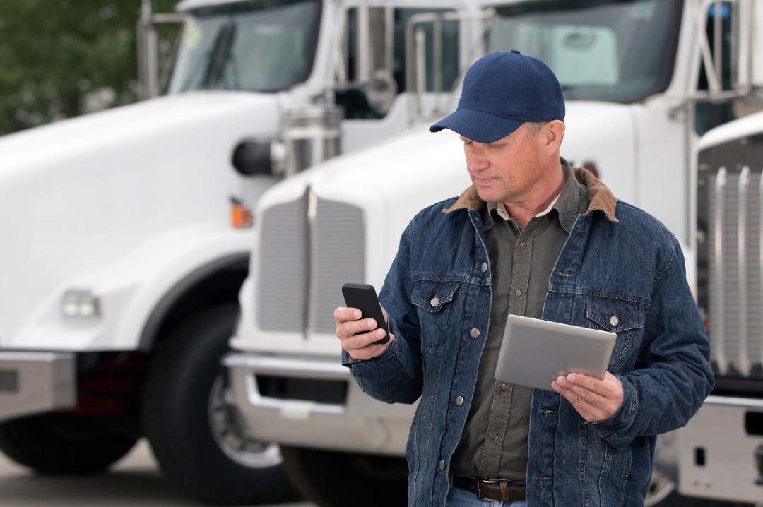 A fleet owner checking his phone and holding a tablet in front of his trucks